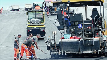 Foto von Asphaltarbeiten, zwei Asphaltbaumaschinen, daneben stehen drei Arbeiter mit Besen und Schaufeln