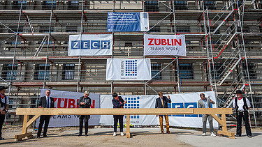 Foto von mehren Personen, die mit einem Hammer Nägel in Holz schlagen. Sie stehen auf der Baustelle der JVA Willich I.