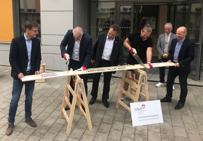 Foto von mehreren Männern im Anzug, die ein Brett sägen. 