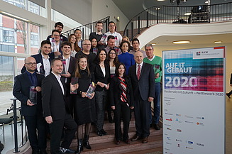 Foto von mehreren Personen auf einer Treppe. Sie sind schick angezogen und halten ein Pokal in den Händen. Neben ihnen ist ein Banner von Bauberufe mit Zukunft – Wettbewerb 2020.     