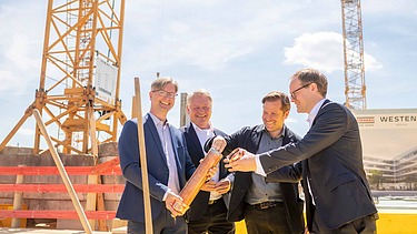 Foto von vier Männern in Anzug auf einer Baustelle. Sie halten eine Röhre. 