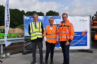 Foto von drei Personen in Schutzkleidung vor einem großen Bauzauntransparent