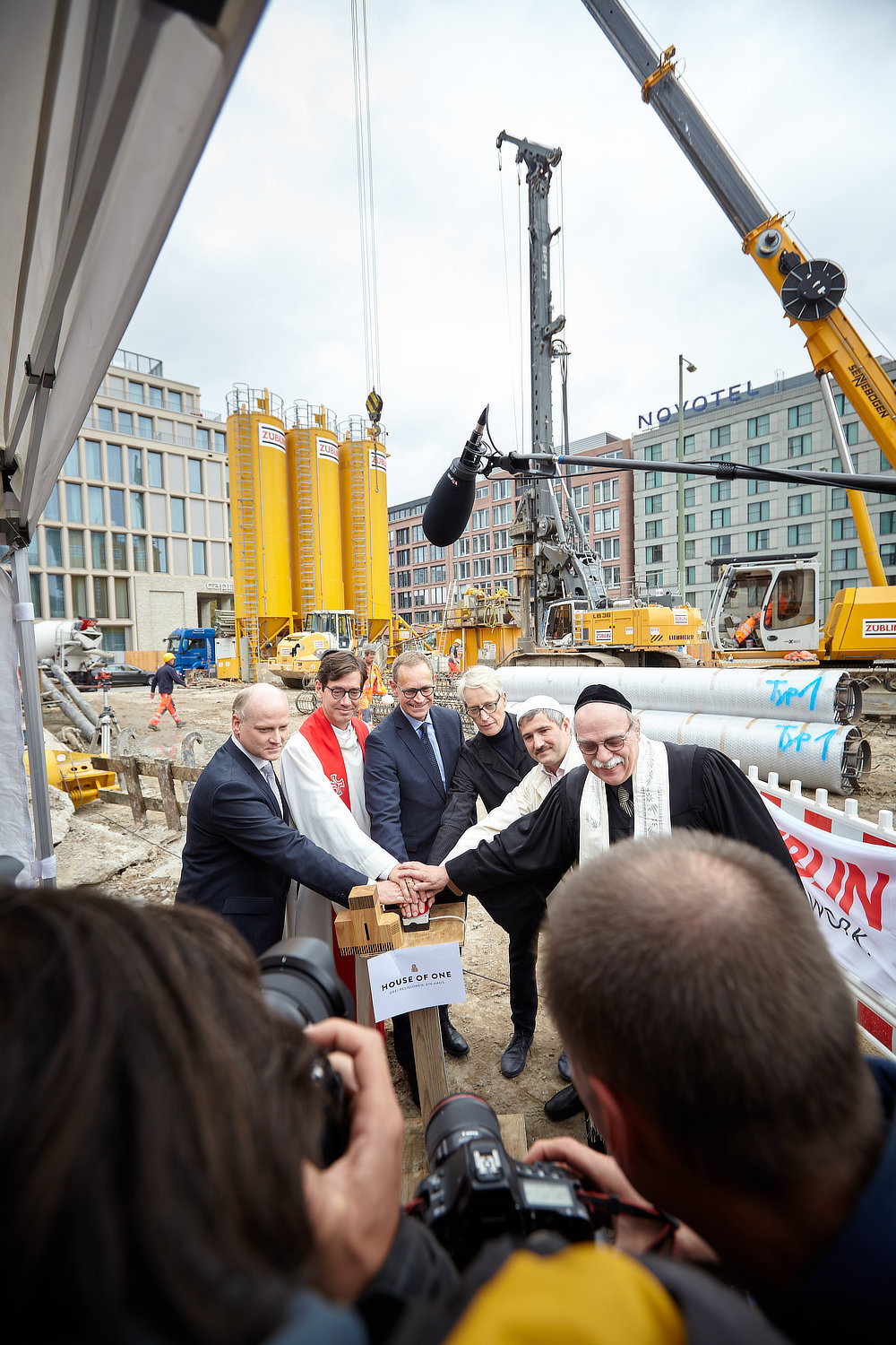 Foto von sechs Männern, darunter drei Priester, auf einer Baustelle, die gemeinsam einen Buzzer drücken. 