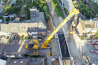 Foto von einem Wohngebiet, wo ein Kransteht der ein Teil für eine Brücke hält.  