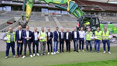 Foto von mehren Personen am Rand eines Fußballplatzes. Hinter ihnen steht ein Bagger. 