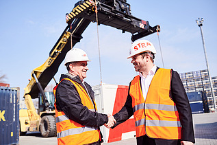 Photo of two people shaking hands and wearing protective clothing, behind which you can see an excavator with its gripper arm extended