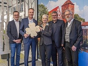 Foto von fünf Personen, die vor einer Bilderwand stehen, die zweite von links hält ein großes Kleeblatt aus Holz in der Hand