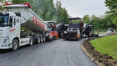 Foto von einem STRABAG LKW beim Abladen, daneben eine Asphaltbaumaschine