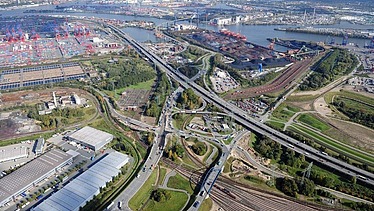 Luftaufnahme von einem Autobahn-Drehkreuz, links steht eine große Lagerhalle, im Hintergrund ist ein Hafen zu sehen