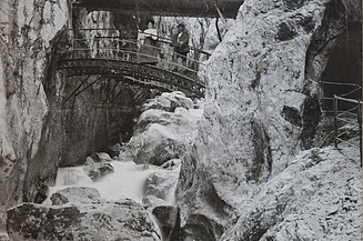 Schwarz-weiß Foto von einer Brücke, die über einen Graben geht. Drumherum sind Bergwände, die sich zu einer Schlucht formen. Auf der Brücke sind zwei Personen zu sehen. 