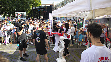 Foto von einem ZÜBLIN-Stand, wo ein Roboter gefilmt wird. Drumherum sind viele Personen versammelt    