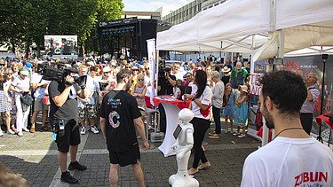 Foto von einem ZÜBLIN-Stand, wo ein Roboter gefilmt wird. Drumherum sind viele Personen versammelt    