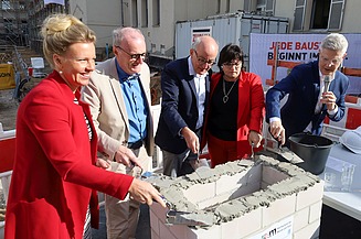 Foto von mehreren Personen, die schick gekleideten sind, die Beton auf ein Gemäuer verstreichen. 