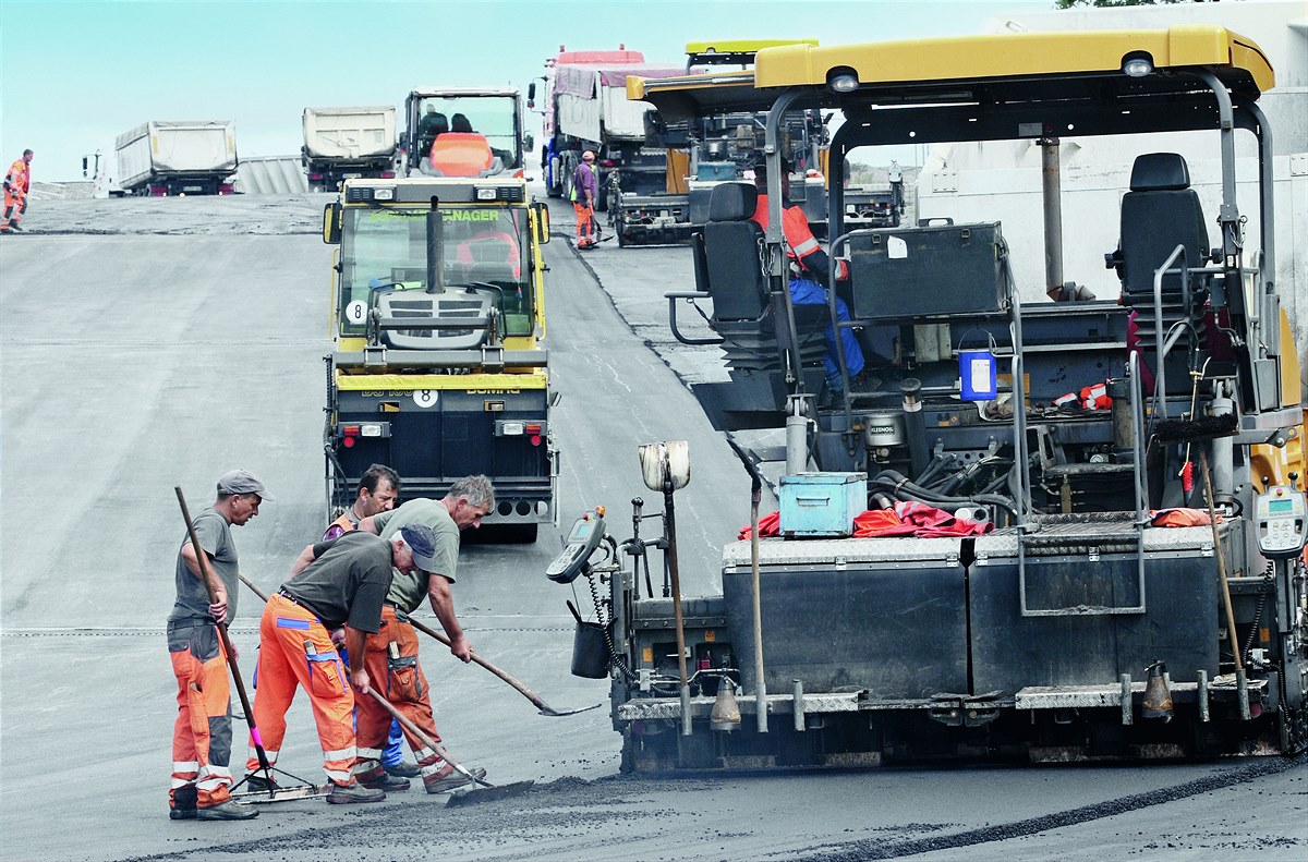 Foto von Asphaltarbeiten, zwei Asphaltbaumaschinen, daneben stehen drei Arbeiter mit Besen und Schaufeln
