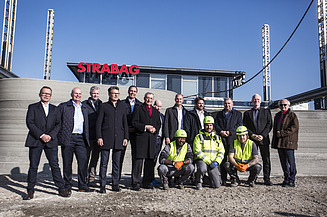 Photo of several people standing in front of a concrete wall, an office building in the background