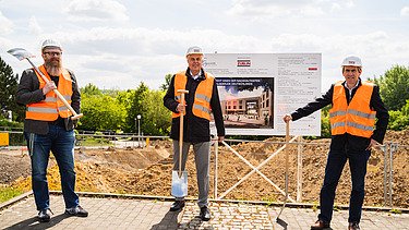 Foto von drei Männern mit Spaten, Wahrfeste und Hell. Hinter ihnen sind Berge aus Erde. 