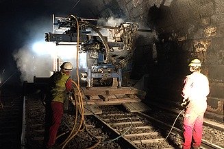 The photo shows two workers with a drilling machine in a tunnel