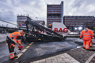 Das Bild zeigt Asphaltierarbeiten mit zwei Arbeitern in Schutzanzügen und einer großen Asphalbaumaschine, im Hintergrund ein Bürogebäude
