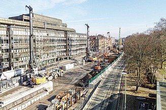 Foto von einer aufgerissenen Straße. Es sind mehrere Kräne und andere Baugeräte zusehen.