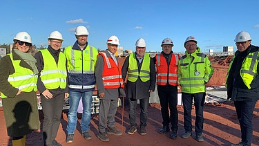 Foto von mehreren Personen die in Warnwesten auf einer Baustelle stehen.  