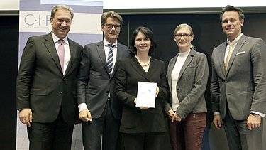 Foto von mehreren, stehenden Personen, die Person in der Mitte hält eine Urkunde in Händen