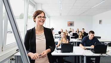 Foto eines Raumes mit mehreren sitzenden Personen, im Vordergrund steht eine weibliche Person mit Brille