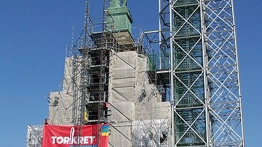  Foto von einer hochgebauten Statue, die auf einem Podest steht. Um das Podest ist ein Gerüst aufgebaut ist.