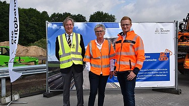 Foto von drei Personen in Schutzkleidung vor einem großen Bauzauntransparent