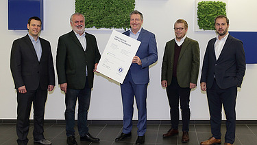 Photo of five people standing, the person in the middle holding a white board in his hand