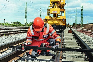 Foto eines Schienenarbeiters in Schutzkleidung, dahinter ein gelbes Schienenbaufahrzeug