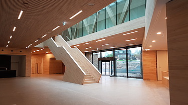 Foto vom inneren eines Gebäudes in der Eingangshalle. In der Mitte erstreckt sich eine Treppe aus Holz.  