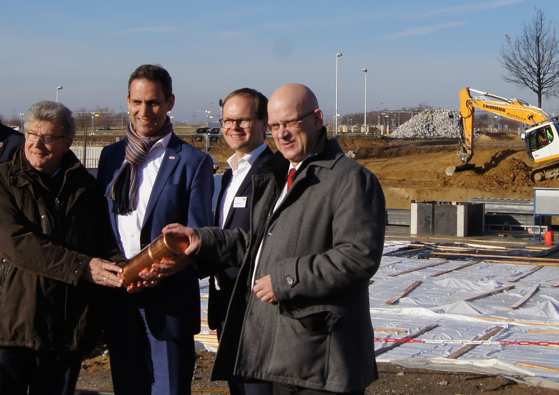 Vier Männer auf der Baustelle in Schönfeld mit symbolischer Grundsteinlegung. Sie halten ein Gefäß.