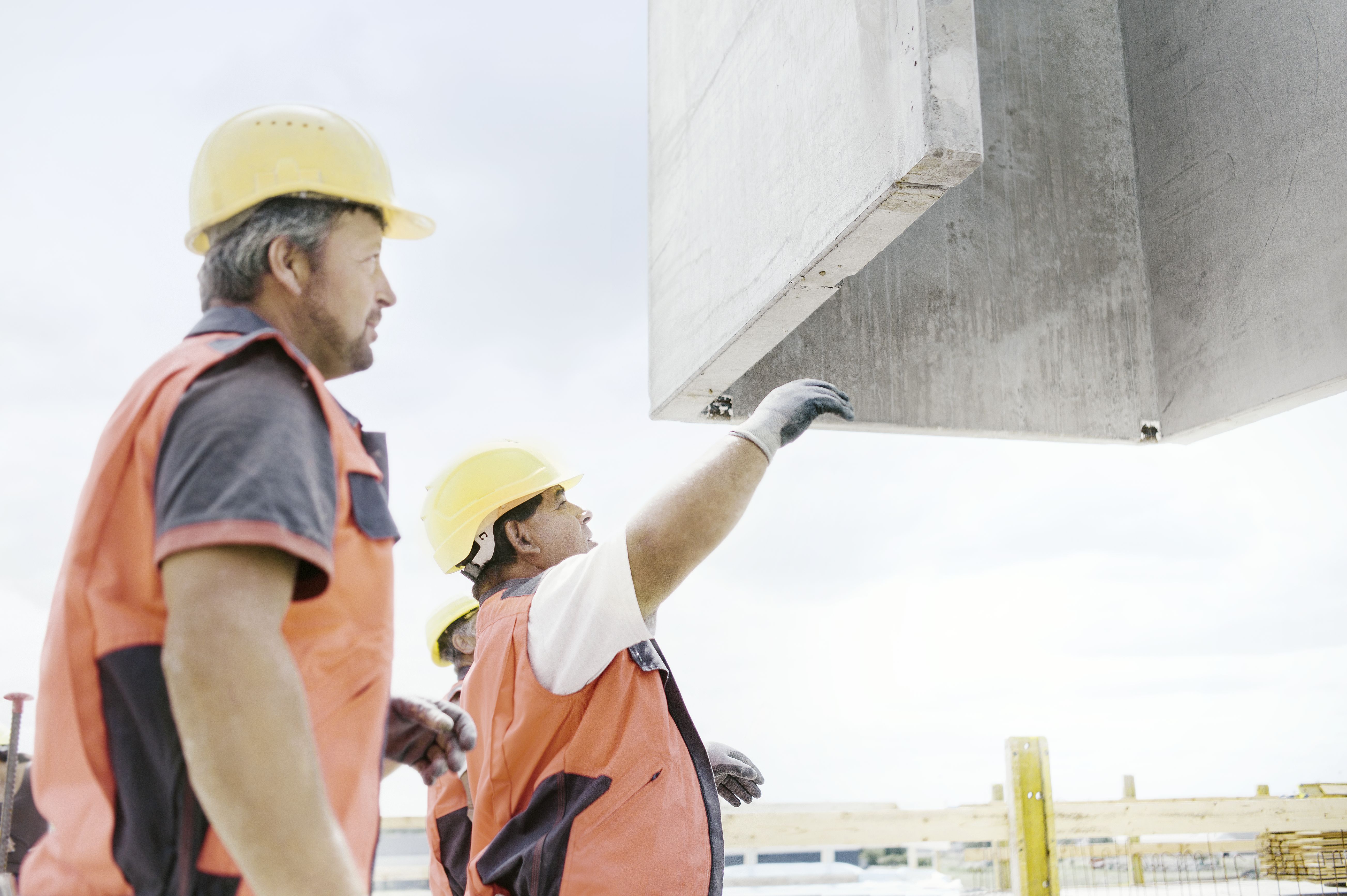 Foto von zwei Personen, die in Arbeitsuniform einen Betonblock navigieren, dass er nach oben gezogen wird. 