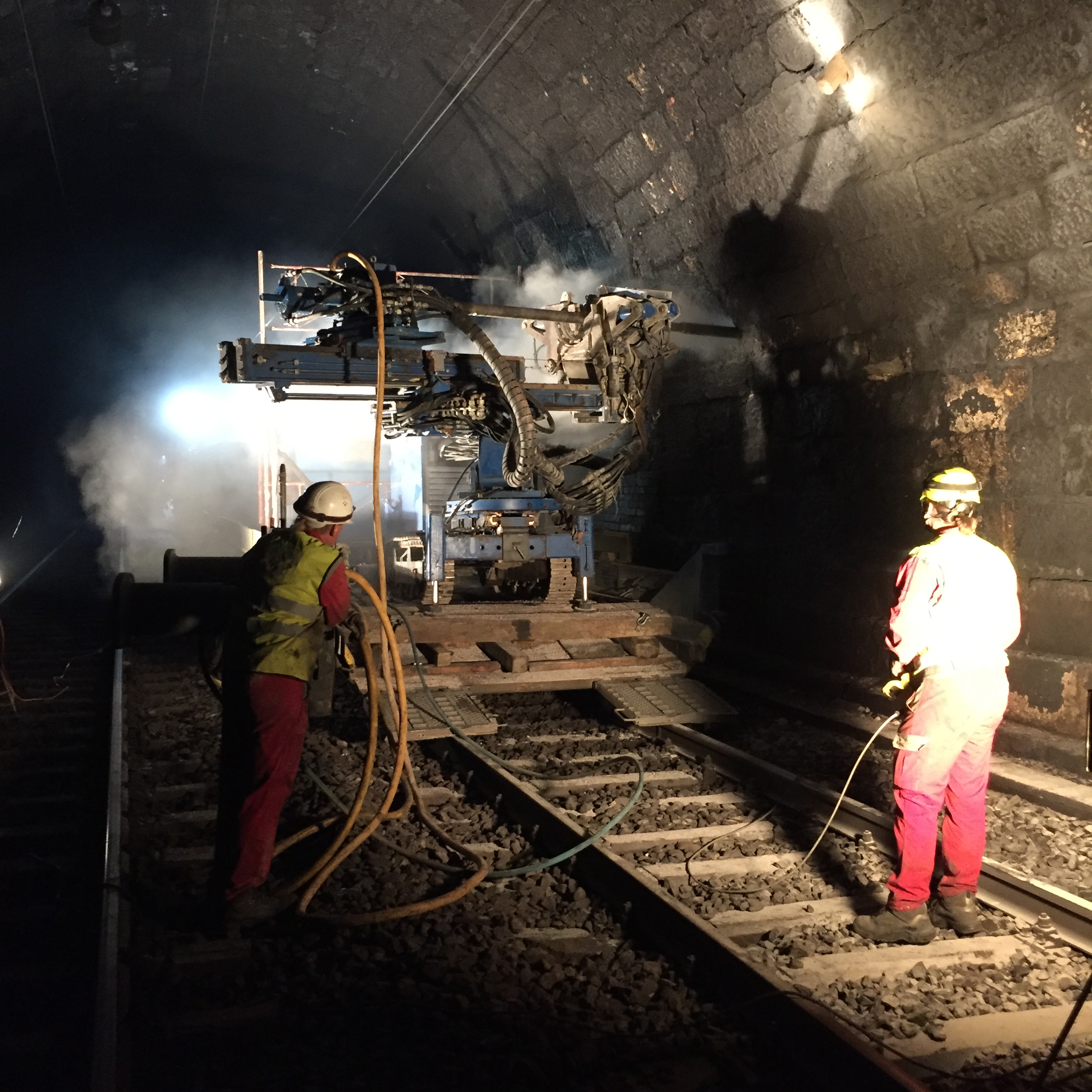The photo shows two workers with a drilling machine in a tunnel