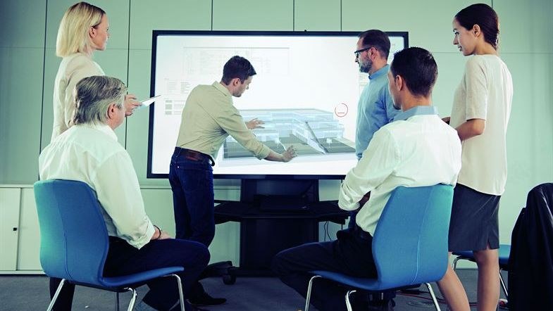 Foto von mehren Personen, die vor einem Fernseher stehen. Es ist eine Präsentation von dem Modele eines Hauses zusehen. Eine Personen verdeutlicht etwas bei der Präsentation.         