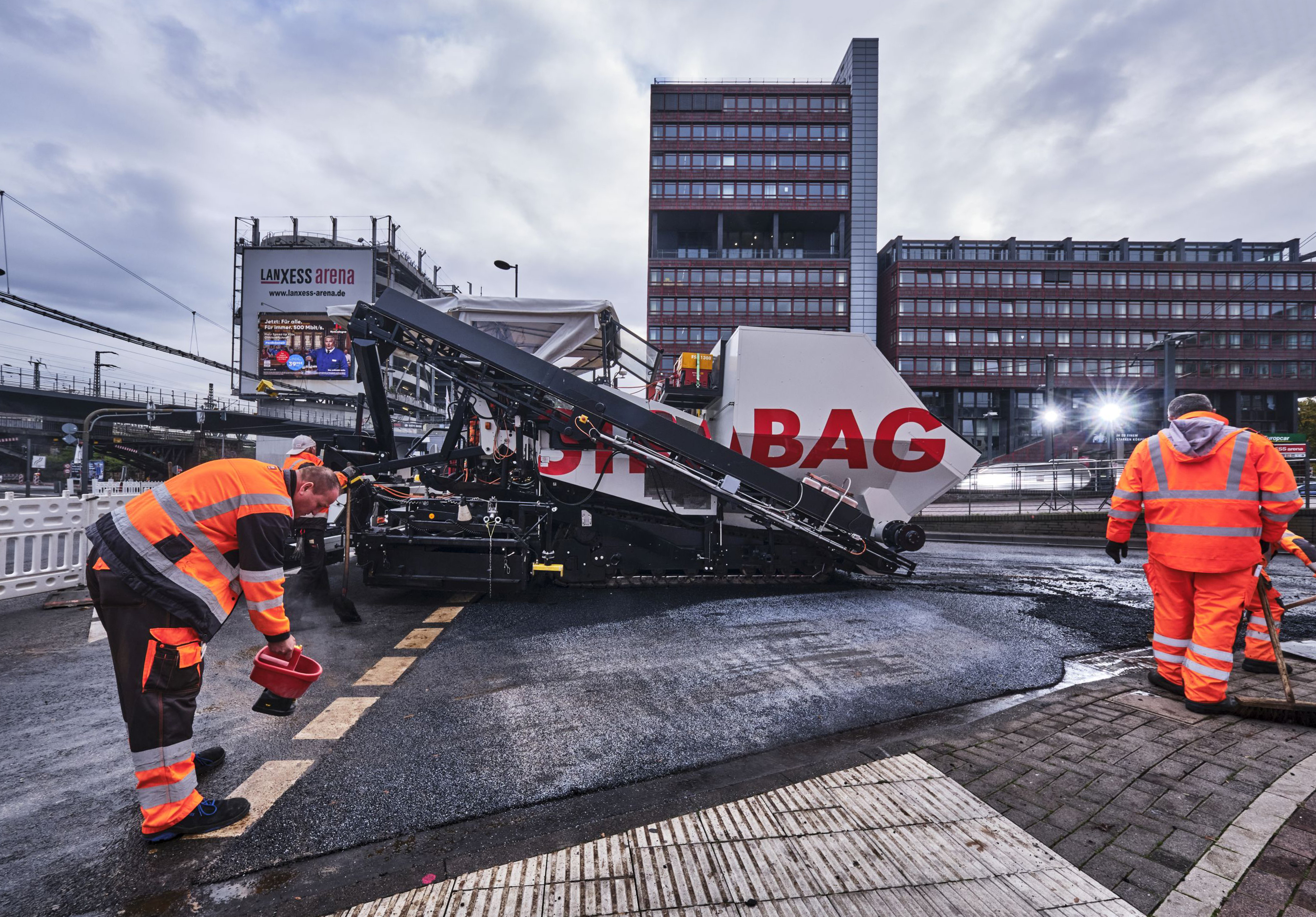 Das Bild zeigt Asphaltierarbeiten mit zwei Arbeitern in Schutzanzügen und einer großen Asphalbaumaschine, im Hintergrund ein Bürogebäude