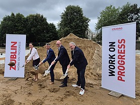 The picture shows four people with shovels at the ground-breaking ceremony, with two company roll-ups to the left and right