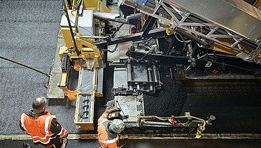 Foto zeigt zwei Straßenbauarbeiter in Schutzwesten vor einer Asphaltbaumaschine