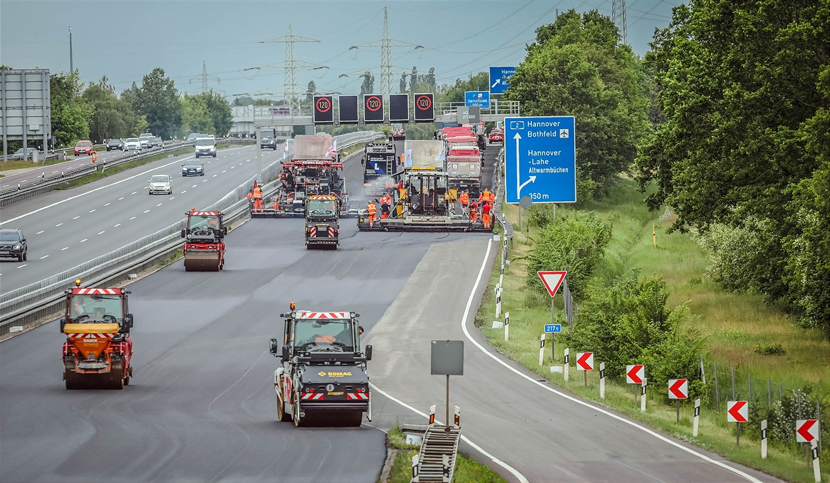 Foto einer Autobahnbaustelle mit mehreren Straßenbaumaschinen darauf, rechts ein blaues Verkehrsschild