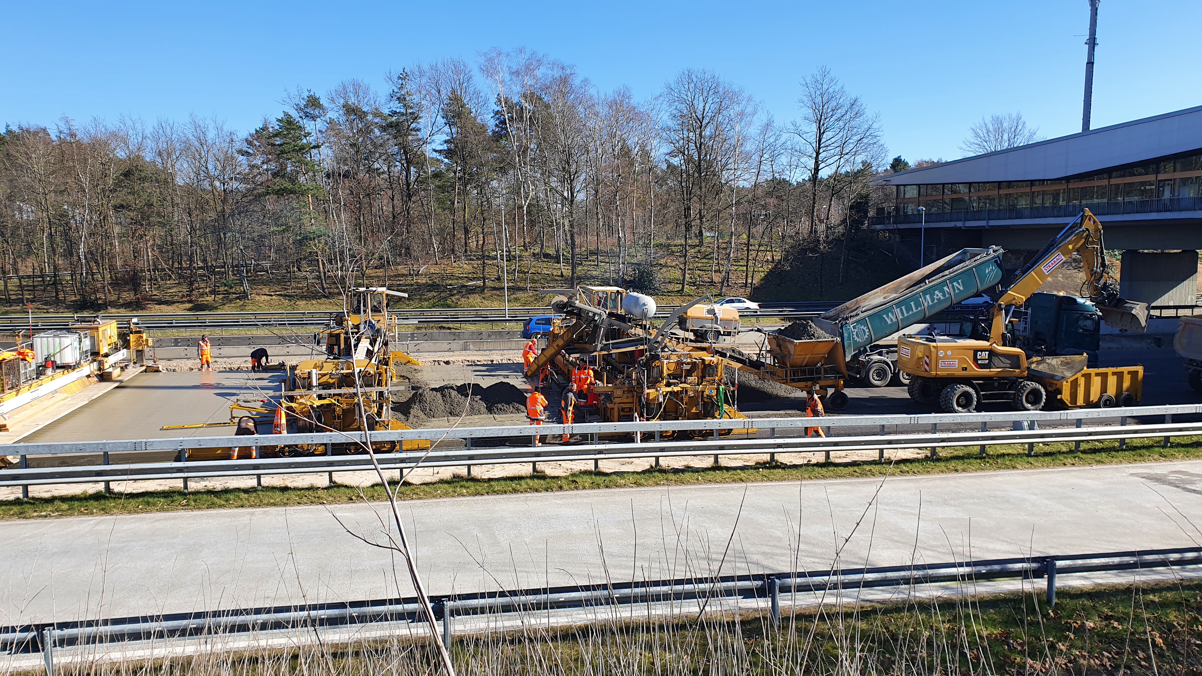 Das Bild zeigt die seitliche Ansicht von mehreren Straßenbaumaschinen beim Einbau einer Betondecke
