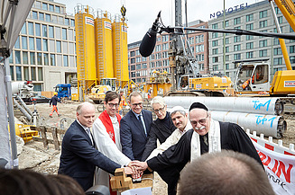 Foto von sechs Männern, darunter drei Priester, auf einer Baustelle, die gemeinsam einen Buzzer drücken. 