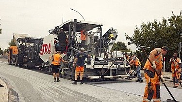 Foto von mehreren Straßenbauarbeitern rund um eine große Asphaltbaumaschine