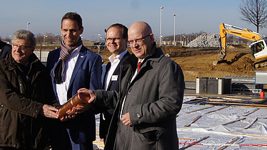 Vier Männer auf der Baustelle in Schönfeld mit symbolischer Grundsteinlegung. Sie halten ein Gefäß.