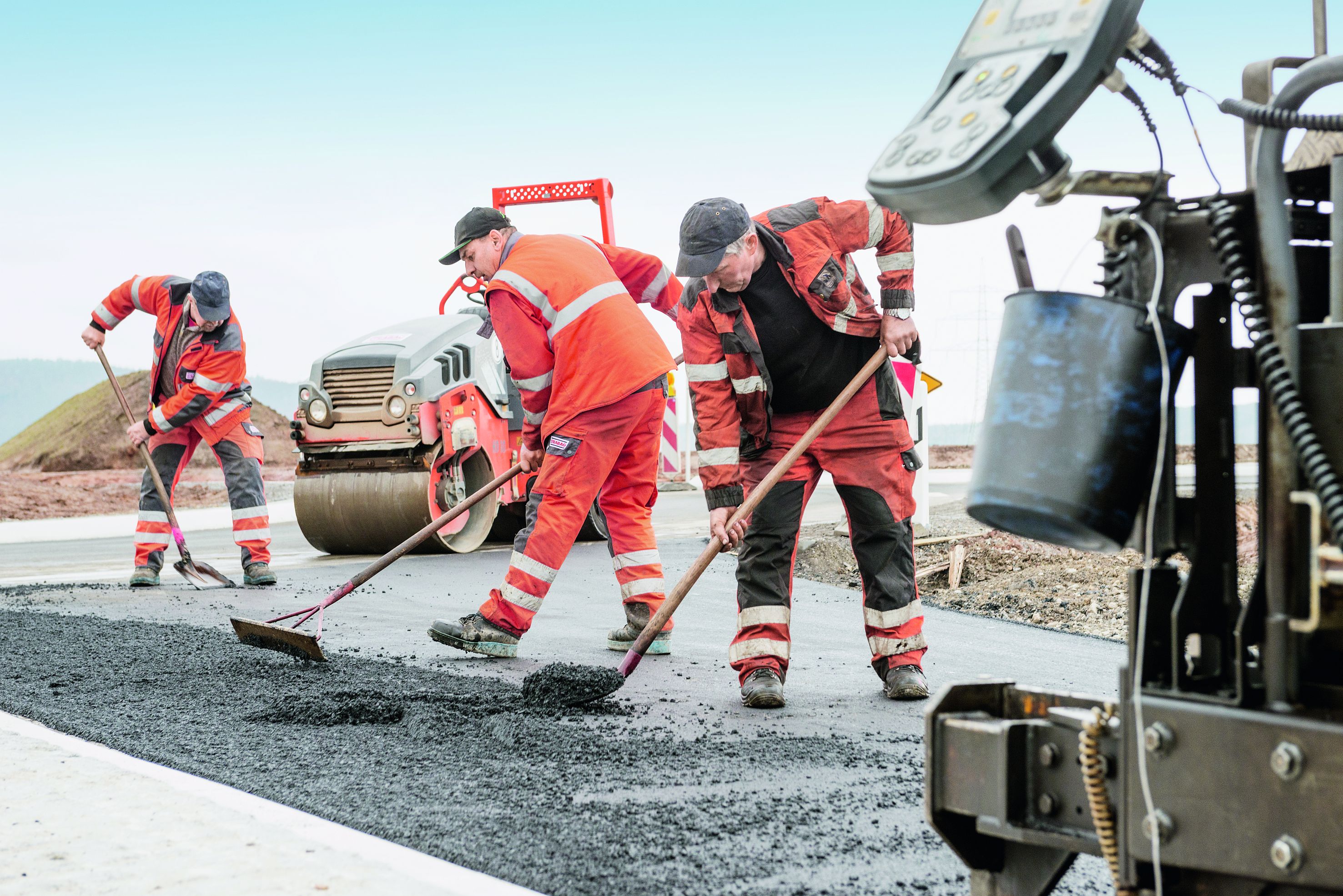 Foto von drei Straßenbauarbeitern mit Besen bei der Arbeit, rechts im Vordergrund sieht man einen Teil einer Straßenbaumaschine
