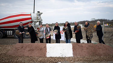 Foto von mehreren Personen beim symbolischen Spatenstich, links hinten steht ein LKW Betonmischer, der rot-weiß gestreift sit