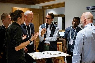 Foto von mehreren Personen im Gespräch, die rund um einen kleinen Tisch stehen