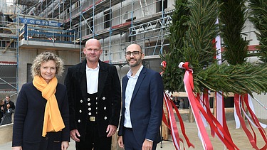 Foto von drei freundlich schauenden Personen auf einer Baustelle. Sie stehen vor dem Richtkranz.  