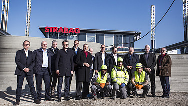 Photo of several people standing in front of a concrete wall, an office building in the background