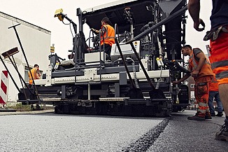 Foto von einer Asphaltmaschine bei der Arbeit, eine Person sitzt in der Maschine, links und rechts stehen noch drei weitere Straßenbauarbeiter