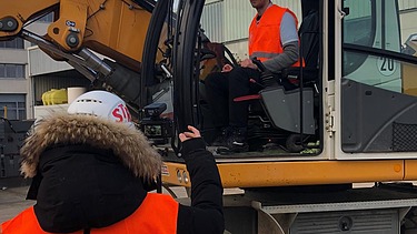 Foto von zwei Personen, die eine sitzt in einem Bagger, die andere steht davor
